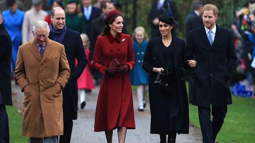 King Charles in a tan coat walks in front of Prince William and next to Kate Middleton in a red jacket, next to Meghan Markle and Prince Harry