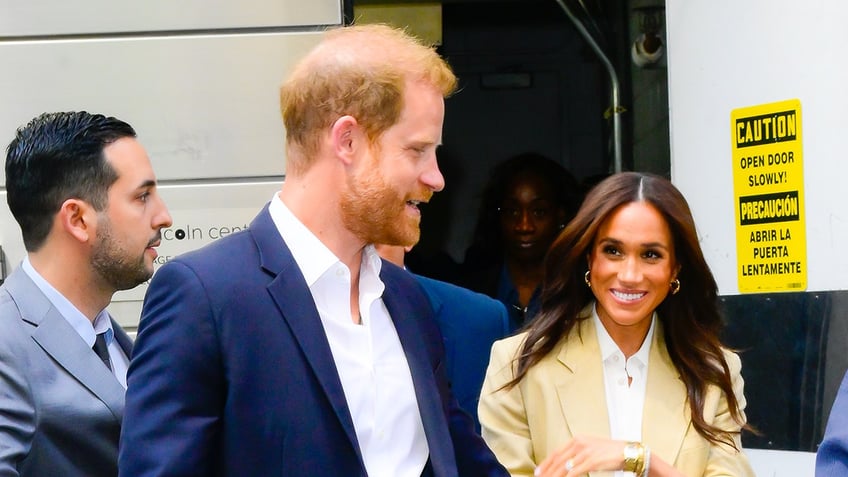 Prince Harry in a blue blazer and white shirt walking next to Meghan Markle in a beige jacket and white shirt.