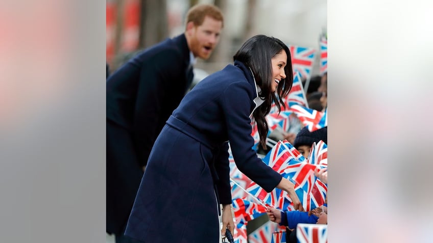 Meghan Markle greeting a crowd holding Union Jack flags as Prince Harry looks on.