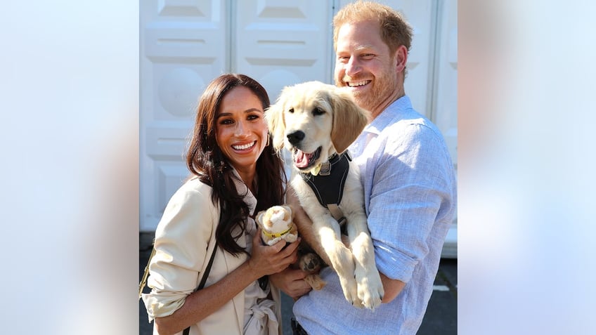 Prince Harry holding a dog as Meghan Markle smiles and poses next to it.