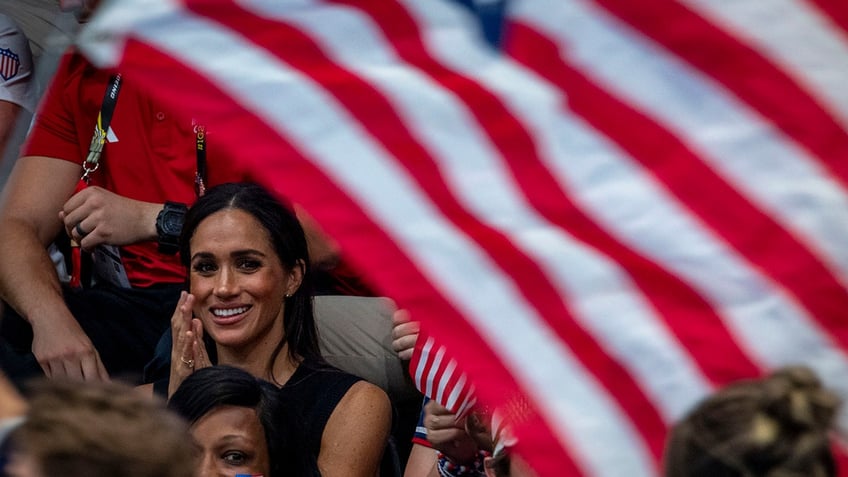 Meghan Markle sitting and smiling as an American flag waves near her.