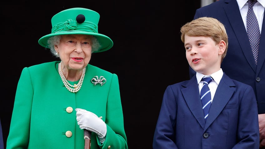 Prince George in a blue suit speaking to his great-grandmother, Queen Elizabeth, in a green suit with a matching hat.