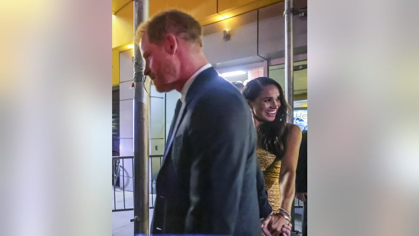 Meghan Markle smiling outisde NYC at night as she holds a serious Prince Harrys hand