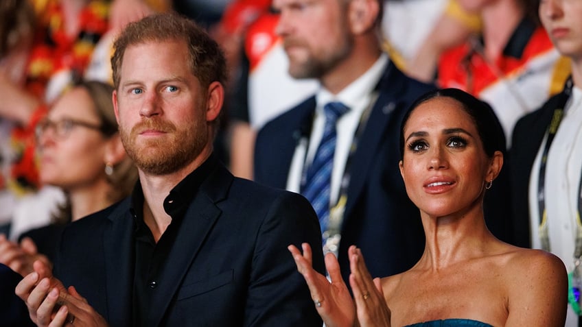 Meghan Markle and Prince Harry looking serious while applauding at the center of a crowd