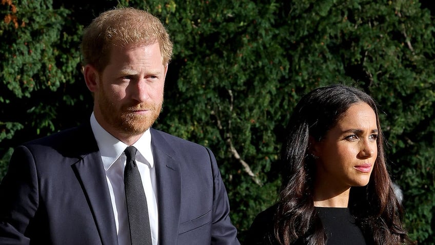 Prince Harry wearing a navy suit and Meghan Markle wearing a black dress as they look serious.