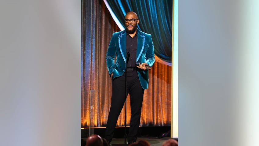 Tyler Perry wearing a bright blue velvet coat and a dark suit speaking on stage.