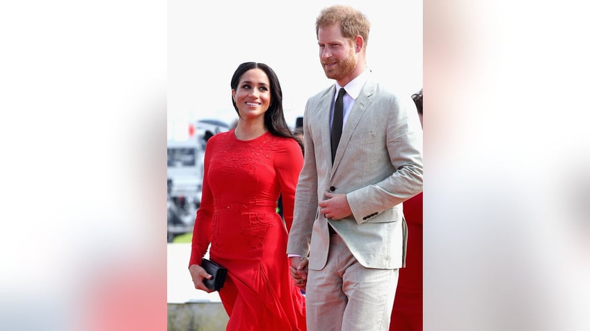 Meghan Markle wearing a red dress smiling at Prince Harry wearing a grey suit and dark tie.