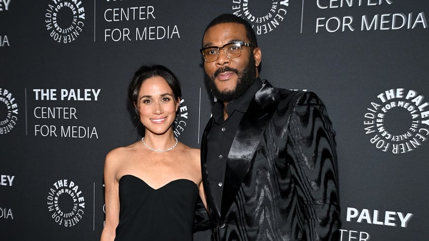 Meghan Markle wearing a strapless sweetheart cut black dress standing next to Tyler Perry wearing all black.