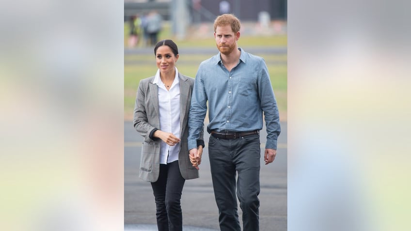 Meghan Markle wearing a white blouse and a grey jacket walking alongside Prince Harry wearing a blue shirt and dark blue pants.