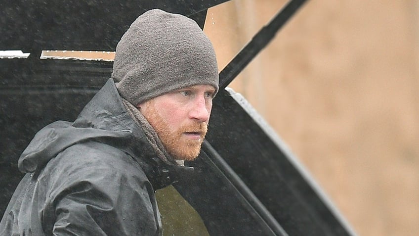 Prince Harry looking to the side wearing a grey windbreaker and a matching hat.