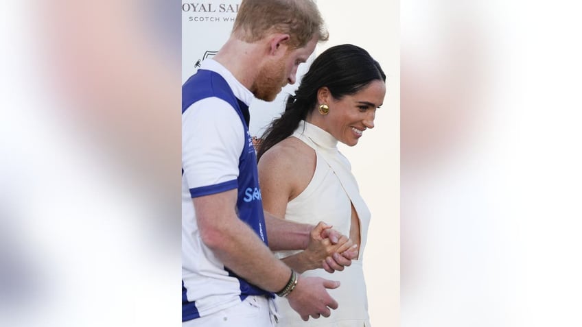 Prince Harry in a polo suit holding Meghan Markle's hand as she wears a cut-out white dress.