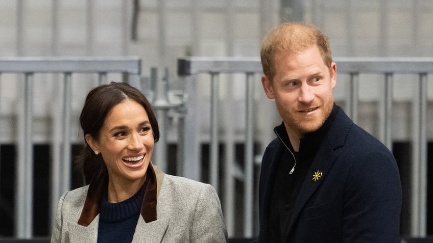 Meghan Markle wearing a beige blazer and a black shirt smiling next to Prince Harry wearing a dark sweater.