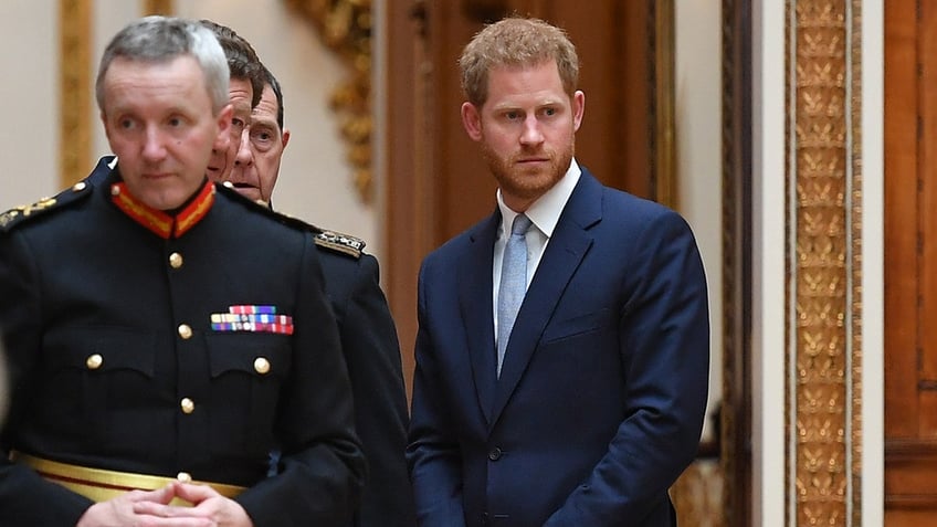 Prince Harry in a navy blazer, white shirt and light blue tie with his hands together next to several men in uniform.