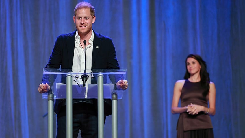 Prince Harry speaking at the podium with an open button white shirt and a dark blazer as Meghan Markle smiles and looks on from a distance.