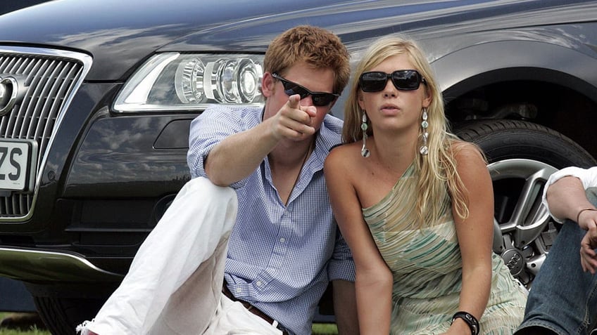 prince harry pointing at the camera as he sits next to Chelsy Davy.