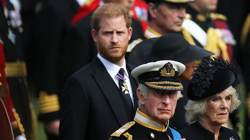 Prince Harry in a dark suit look away as King Charles and Queen Camilla stand in front