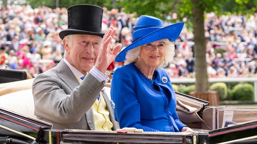 King Charles in a grey suit and black top hat in a carriage next to Queen Camilla wearing a blue coat dress and matching hat.