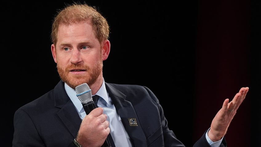 Prince Harry speaking to a mic wearing a dark suit and a light blue shirt.