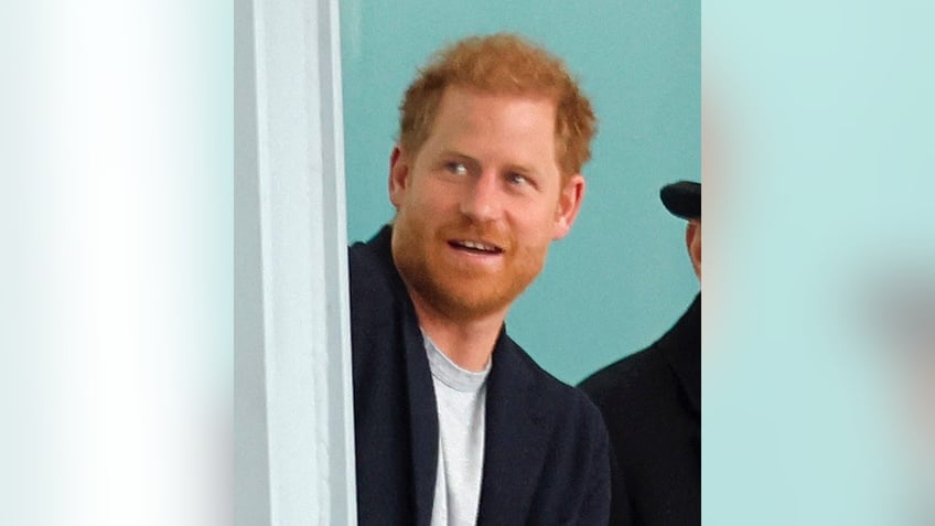 Prince Harry looks up and to his right wearing a blue jacket at Heathrow Airport as he leaves the UK