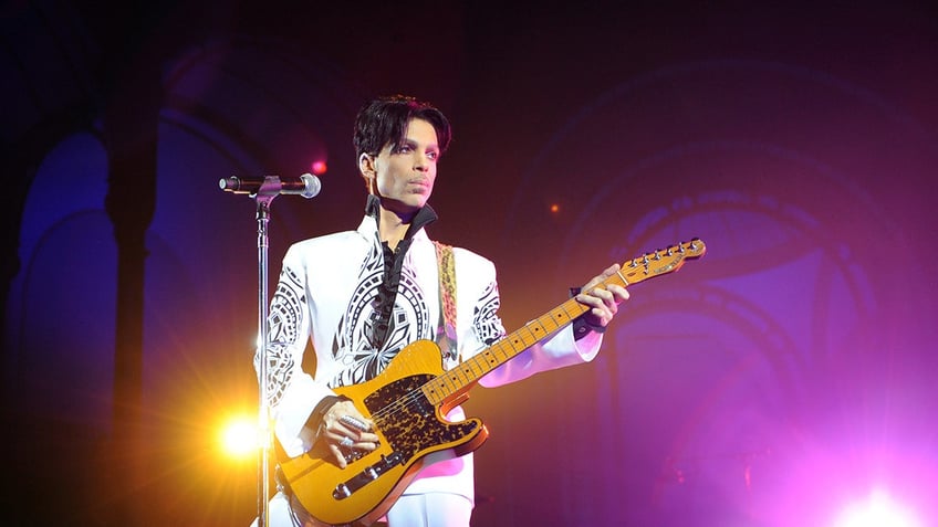 Prince holding a gold guitar on stage wearing a white suit.