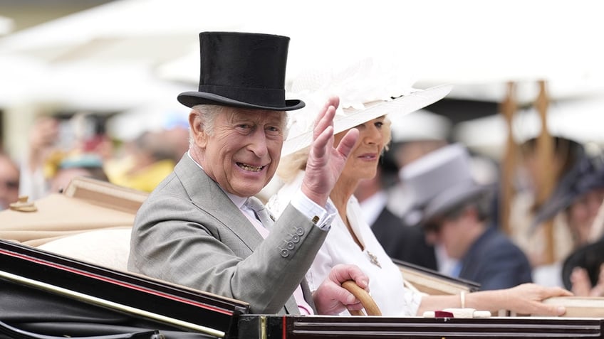 King Charles in a grey suit and black top hat waving in a royal carriage next to Camilla wearing all white with a matching hat.