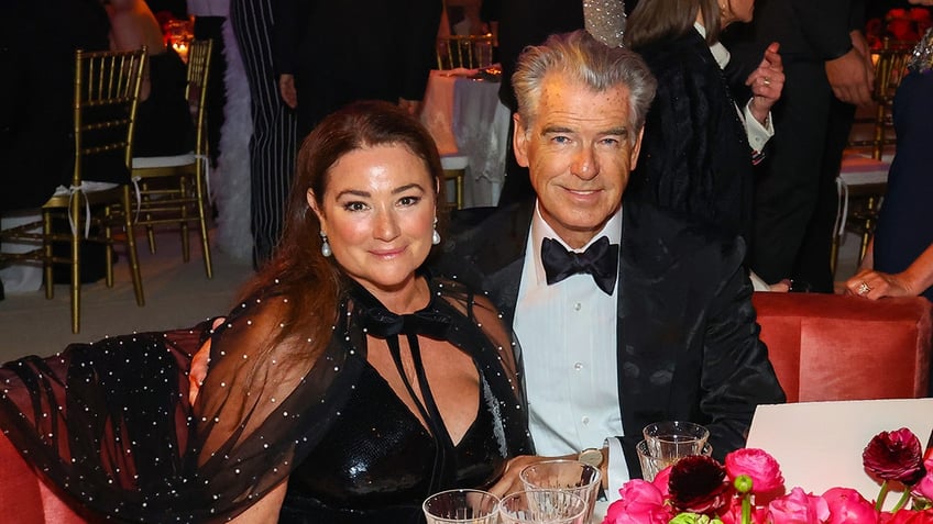 Keely Shaye in a black dress with a mesh shoulder cape sits next to Pierce Brosnan in a traditional tuxedo