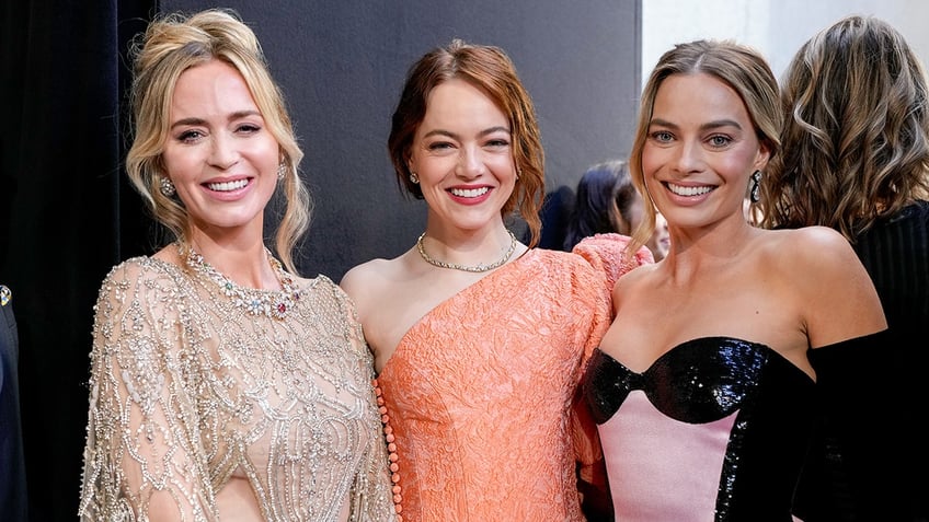 Emily Blunt in a sparkly cut out dress stands next to Emma Stone in an orange sparkly dress who stands next to Margot Robbie in a black and pink dress