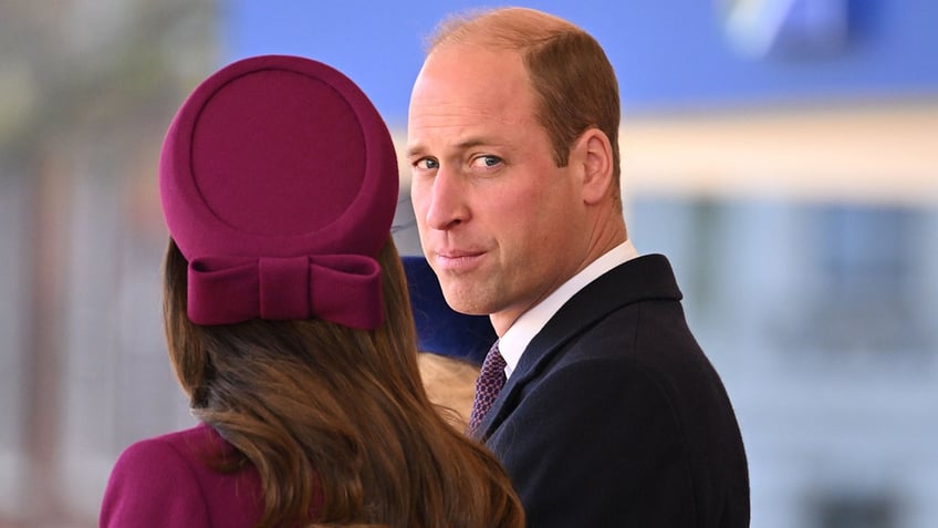 Prince William looks at the camera as Kate Middleton in a maroon outfit has her body faced away from the camera