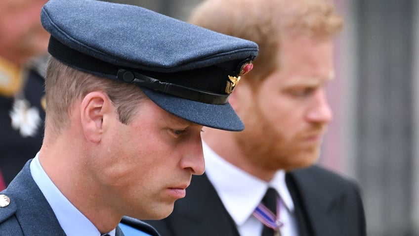 Prince William in uniform standing next to Prince Harry in a suit and looking down.