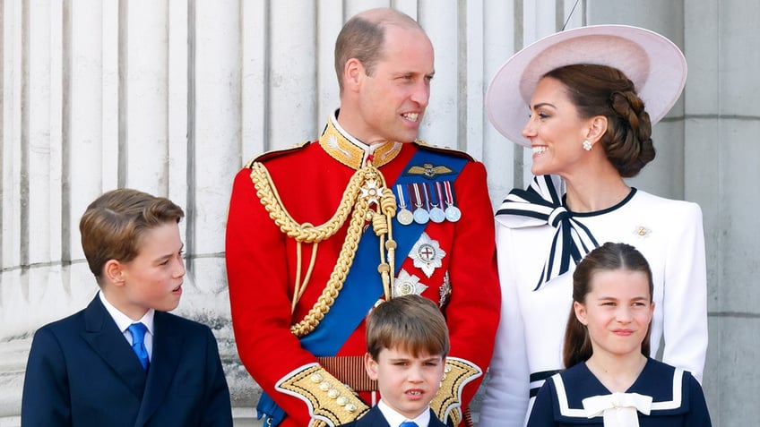 The Prince and Princess of Wales smiling and admiring each other on the palace balcony with their three children.
