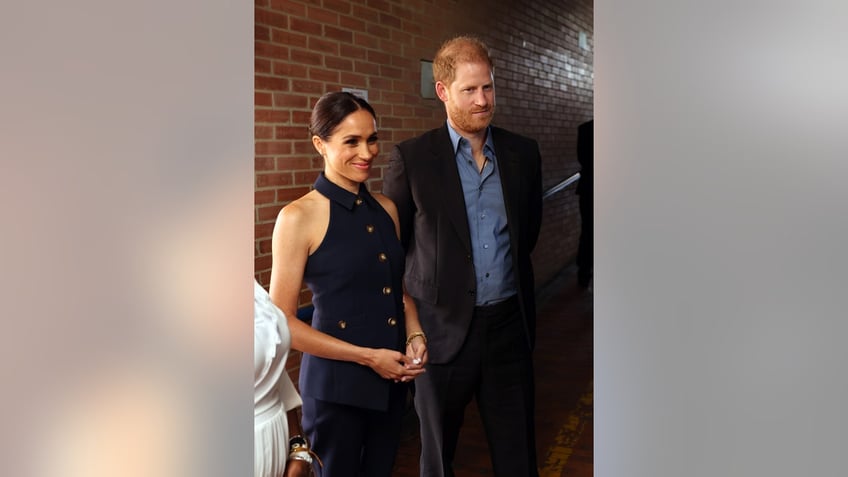 Meghan Markle wearing a sleeveless navy blue dress next to Prince Harry in a dark blazer and blue shirt.