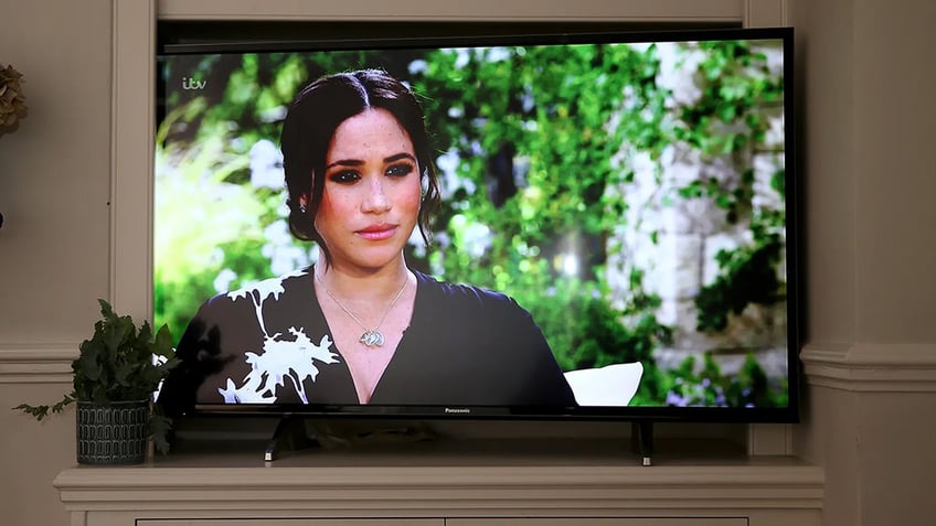 Meghan Markle on a television screen wearing a black and white floral dress against a garden background.