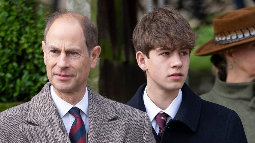 Prince Edward in a brown suit standing in front of his son who is wearing a darker suit.