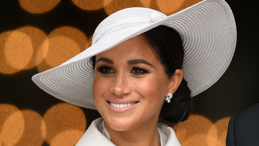 Meghan Markle smiles slightly off-camera in a white hat and matching outfit and earrings