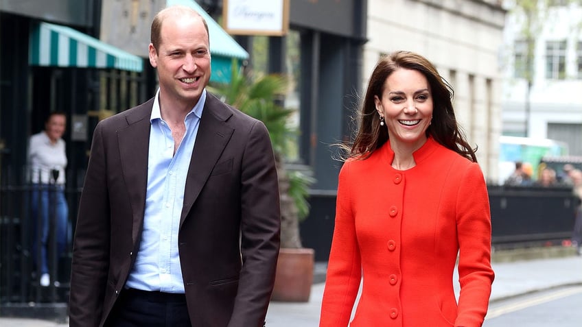 Prince William in a blue shirt and brown coat walks alongside Kate Middleton in a bright red/orange long coat
