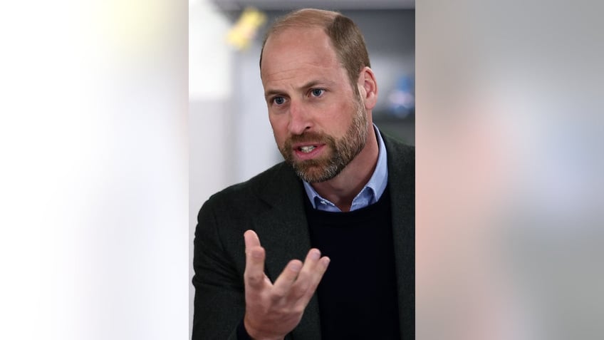 A close-up of Prince William wearing a dark sweater and a light blue collared shirt in conversation.