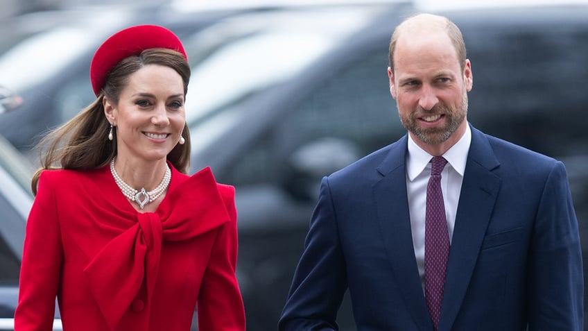 Kate Middleton wearing a red dress and matching hat walking next to Prince William wearing a blue blazer and a purple tie.