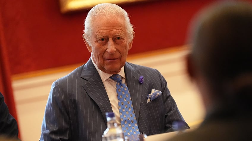 King Charles in a dark blue suit and a light blue printed tie sitting in front of a desk.