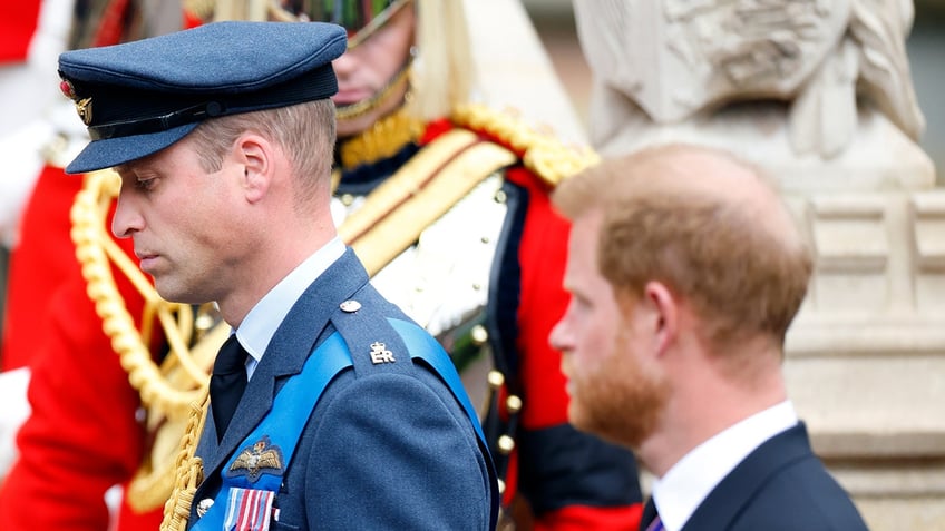 Prince William looking somber in a blue suit with medals walking outdoors ahead of Prince Harry wearing a black morning suit.