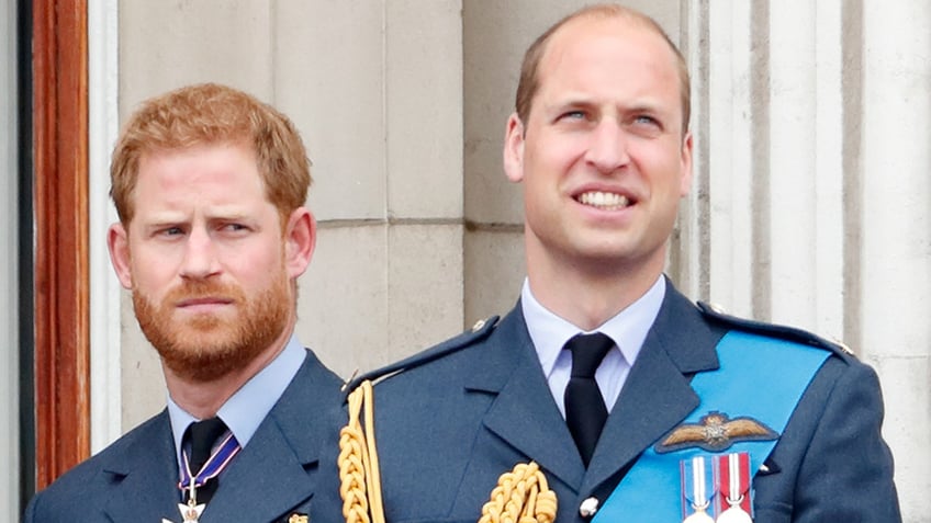 A close-up of Prince Harry and Prince William standing on the balcony of Buckingham Palace wearing matching blue uniforms with medals.