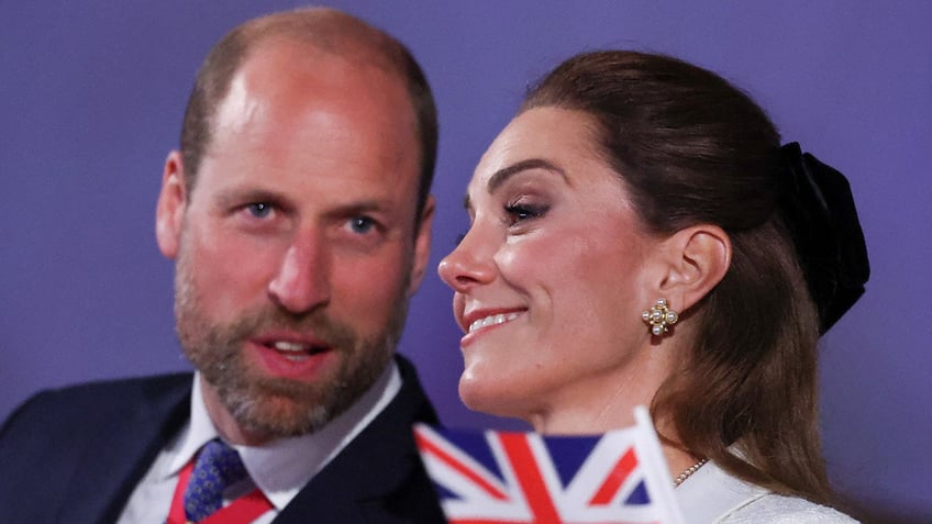 A close-up of Prince William telling something to a smiling Kate Middleton with a Union Jack flag in front of her.