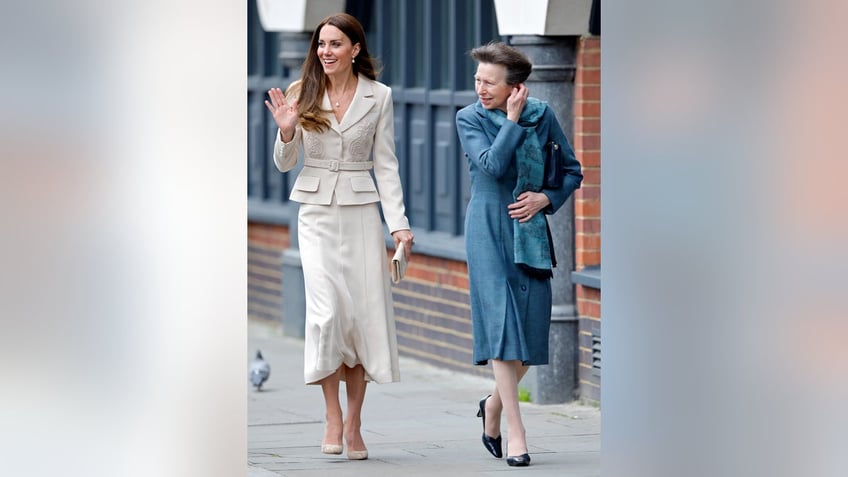 Princess Anne in a blue suit dress walking next to Kate Middleton in a beige suit dress as she waves
