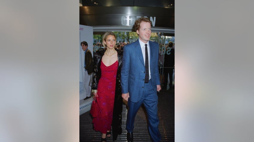 Charles Spencer in a light blue suit and tie walking in front of Caroline Freud in a red dress and black jacket