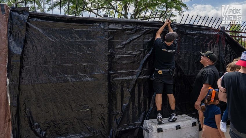 A black tarp is attached to a fence by men in black shirts and shorts