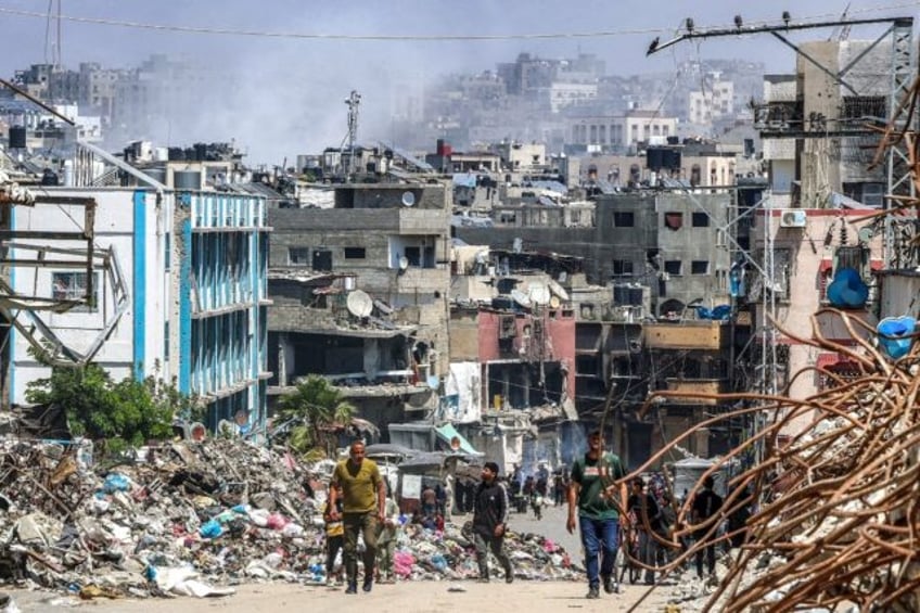 People walk past a mound of trash and destroyed buildings as smoke rises during Israeli bo