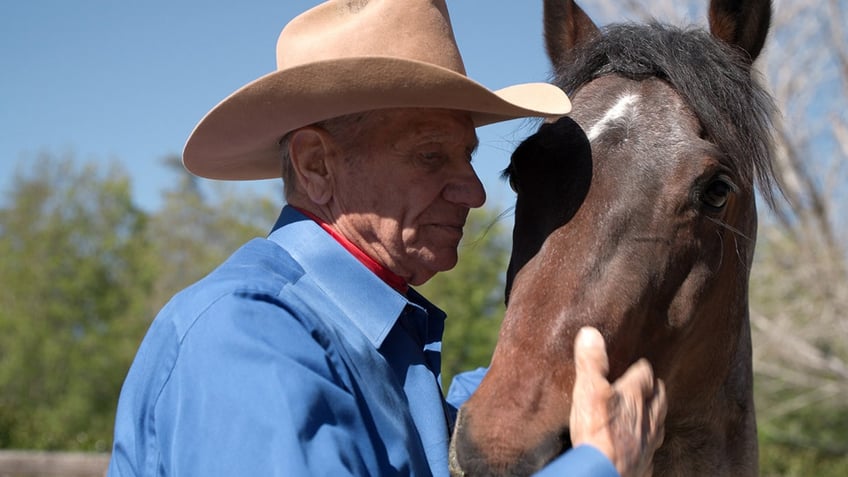 Monty Roberts in a blue shirt and cowboy hat, caressing the head of a brown horse.