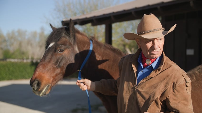 Monty Roberts holding onto a brown horse in cowboy clothes.