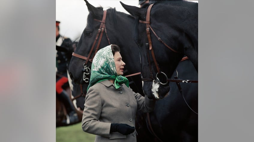 Queen Elizabeth wearing a green scarf and a grey coat surrounded by black horses.