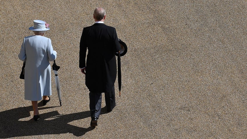 Queen Elizabeth wearing a light blue coat dress and matching hat walking next to Prince Andrew with their backs turned