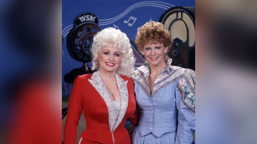 Dolly Parton in a corset red blazer with sparkly silver lapels and Reba McEntire in a blue corset top with patterned sleeves